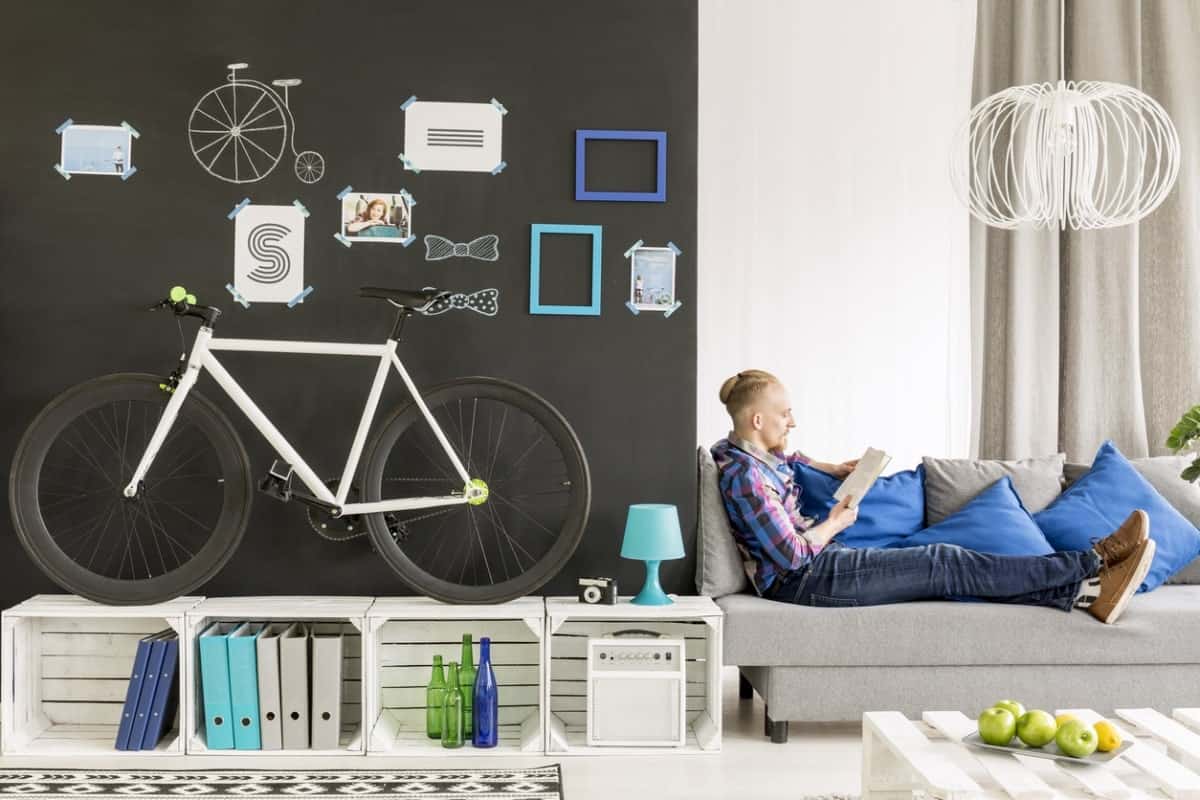一辆自行车陈列在板条箱的架子上，在客厅的黑色墙壁前，一个男人坐在沙发上看书。