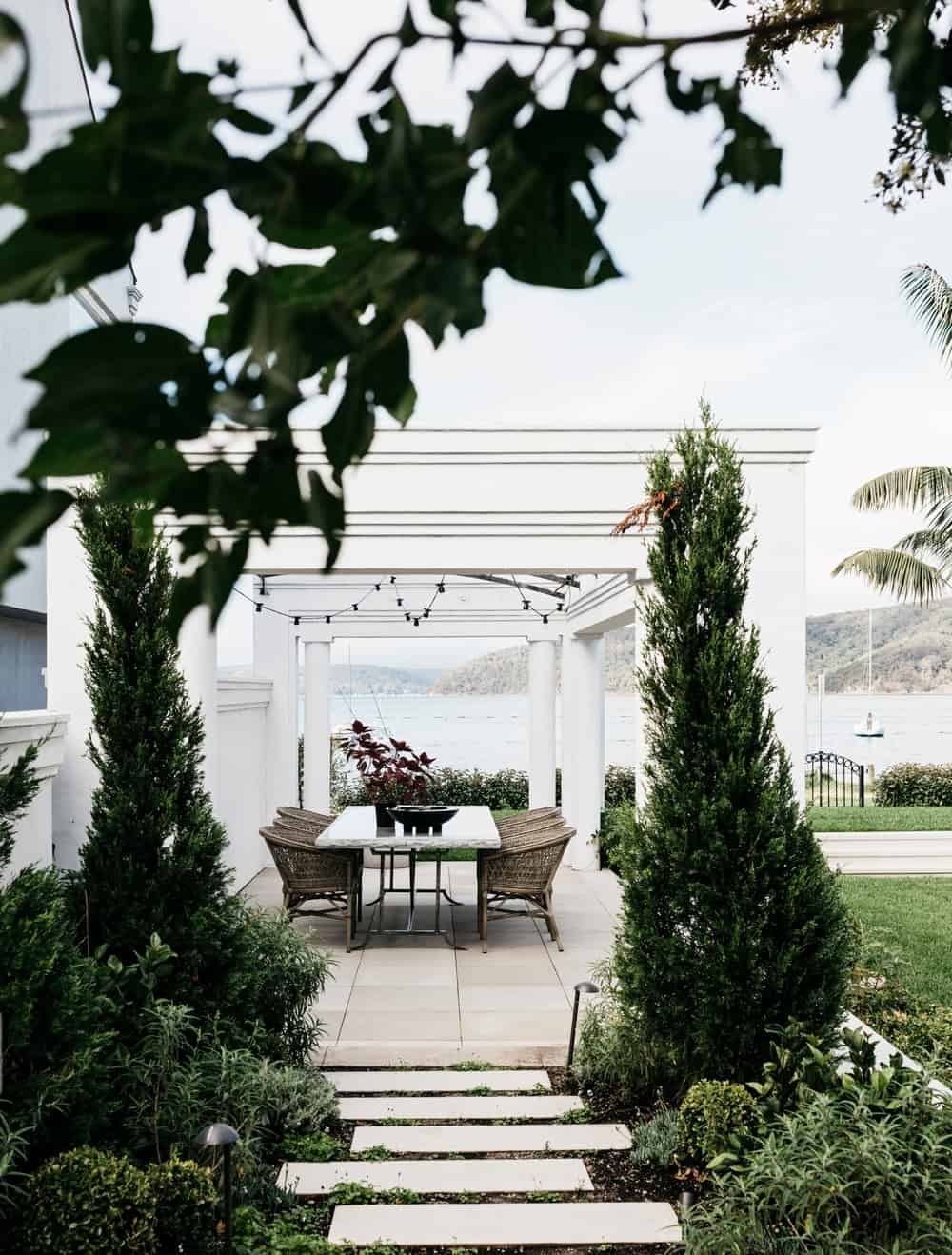 白色的露台上有一条美丽的小径，周围环绕着树木和绿色植物，通往户外餐桌。