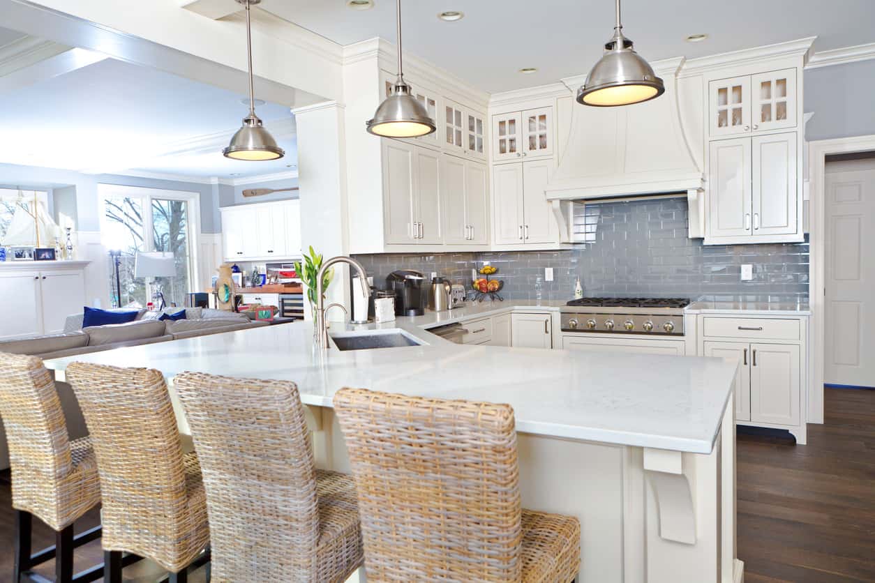 白色厨房，配有镀铬吊灯、蓝色后挡板、深色硬木地板和柳条早餐吧凳。