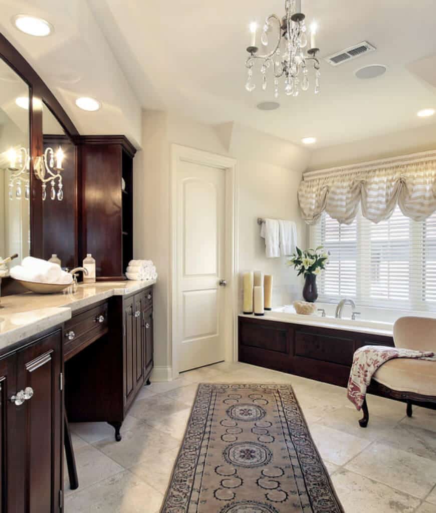 一把米色软垫椅子坐在这个主浴室的浴缸前，有木制洗脸盆和混凝土瓷砖地板，上面有一个带边框的滑道。