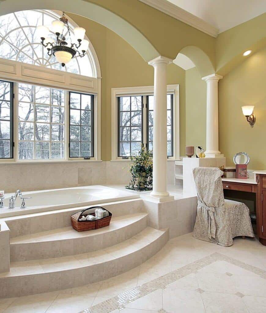自然光通过主浴室的框窗照射进来，主浴室有一个壁凹浴缸，内衬白色柱子。旁边有一个木制的梳妆台和一张有裙摆的椅子搭配在一起。