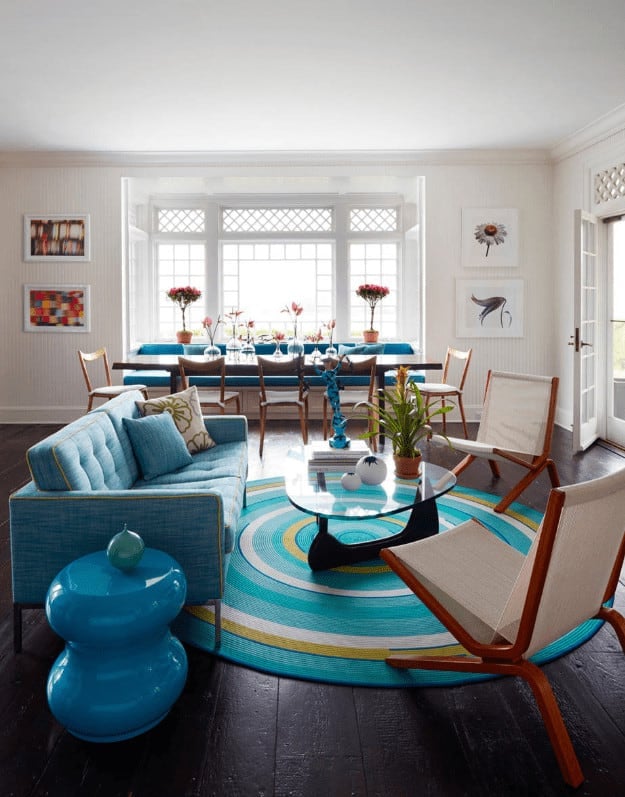 一张玻璃咖啡桌放在一块圆形地毯上，铺在宽大的平板地板上。它配有米色椅子和蓝色簇绒沙发，旁边有一个高光泽光滑的桌子。