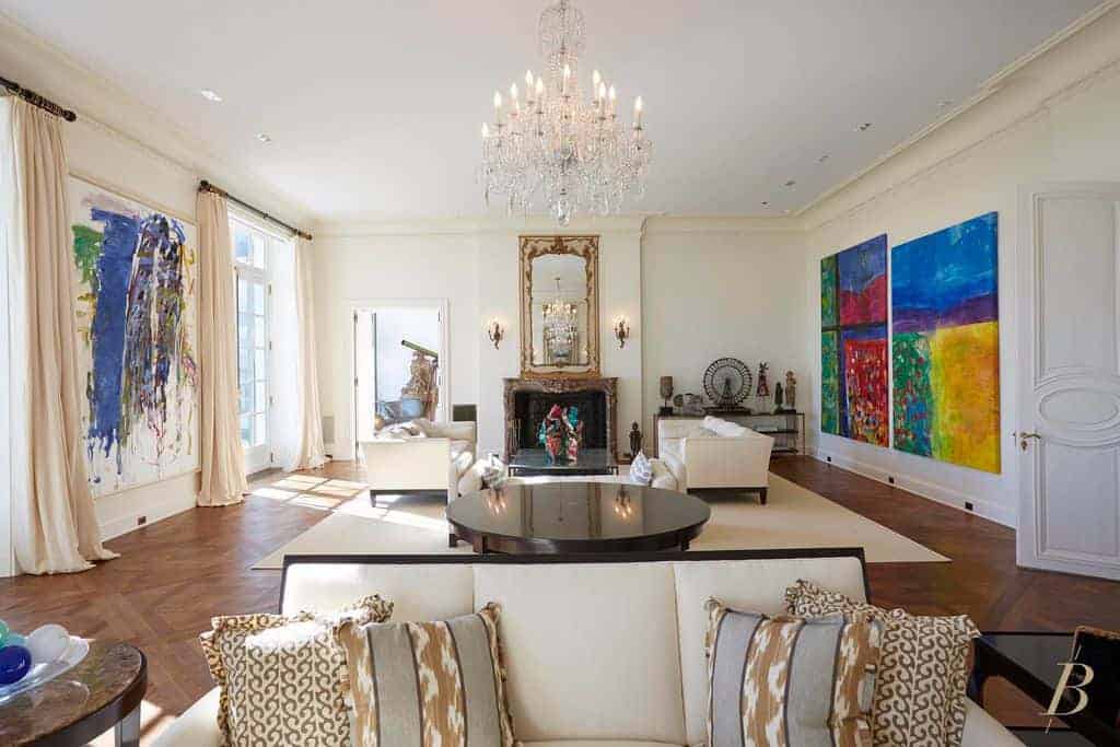 一个别致的水晶吊灯照亮了这个客厅，白色的墙壁上有多个座位和彩色的抽象画。它包括一个壁炉和一面侧面装有烛台的长方镜子。