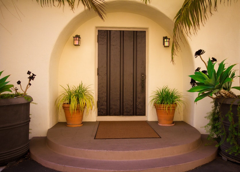 一个简单的木制前门在一个拱形嵌入墙由户外烛台照明。绿色的盆栽植物和混凝土平台上的棕色镶边地毯作为补充。