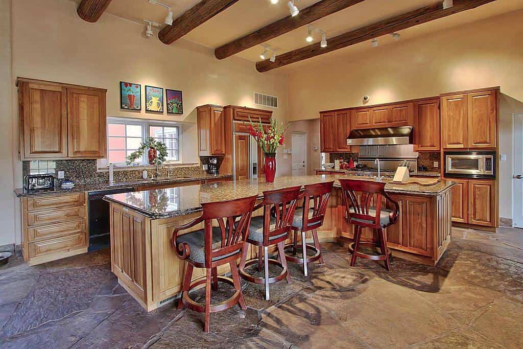 这间可爱的厨房有米色的墙壁和米色的天花板，天花板上有裸露的木质木梁。这与L形厨房岛和半岛上使用的浅色木材以及灰色石头纹理地板和早餐吧的红木凳子非常匹配。