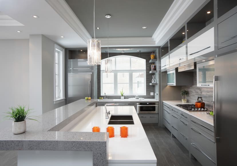 这是一个灰色色调的厨房。它应用于优雅的托盘天花板上，在U形灰色厨房半岛上悬挂着两个水晶吊灯，灰色橱柜和早餐吧上的灰色台面。