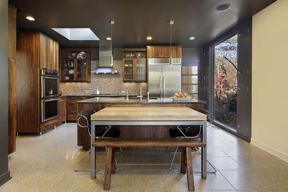这种工业风格的厨房有哑光灰色的墙壁和天花板，带有嵌入式灯，在墙壁的L形结构处的烹饪区域上方有一个天窗。这种结构与餐桌旁的木制厨房岛相匹配，与灰色地砖非常匹配。