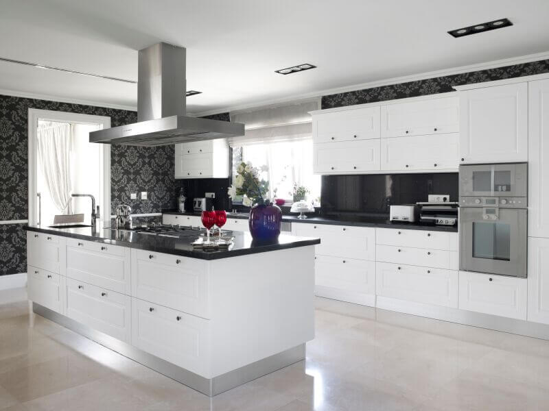 这个厨房里黑白美学的简约是它美丽的主要来源。鲜明的白色橱柜有一个经典的设计，应用在厨房岛和单墙布局。这两者都与黑色花岗岩台面和后挡板以及有图案的黑色壁纸形成对比。