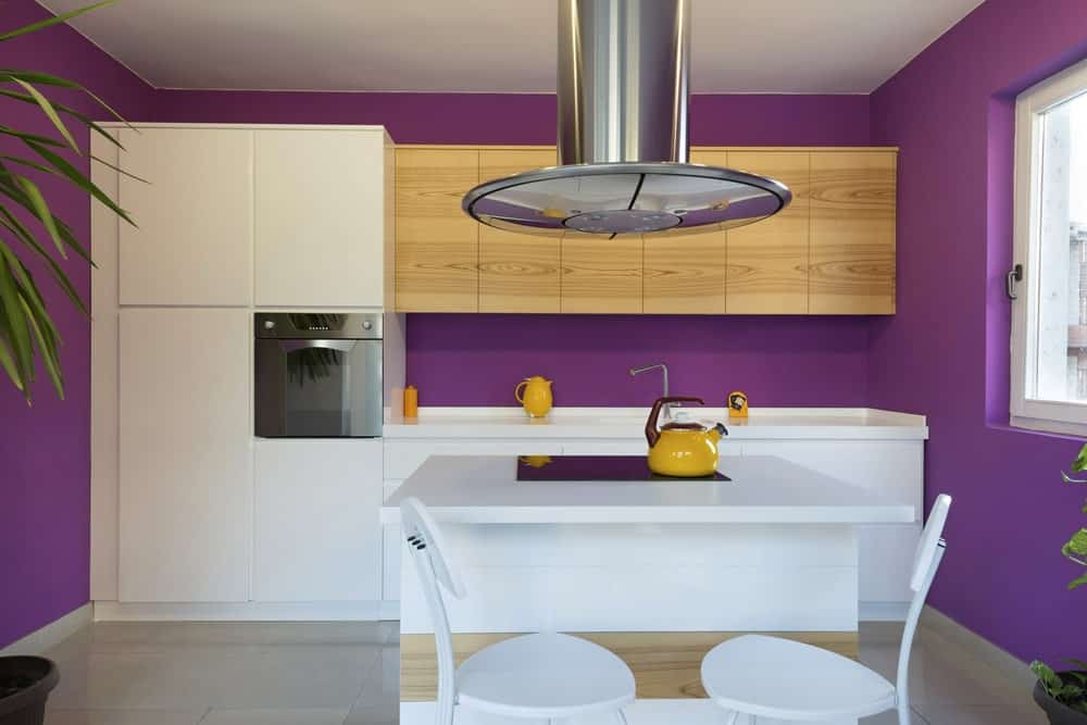 这个小厨房可爱而欢快的紫色墙壁与靠墙的白色现代橱柜相得益彰，水槽上方还有裸露的木制浮动橱柜。这里有一个白色的小厨房岛和一对白色的现代凳子，供早餐吧使用。