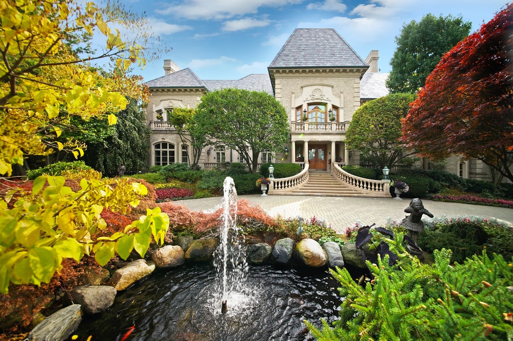 这是房子正面的鸟瞰图，周围有高大的树木、彩色的灌木和一个池塘，池塘的喷泉正对着正门的大庭院。图片来自Toptenrealestatedeals.com。